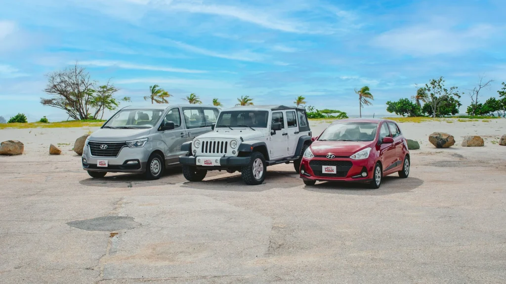 A fleet of three rental cars, a silver minivan, a white Jeep Wrangler, and a red compact car, parked on a parking space with a sunny Aruba beach and blue sky in the background.